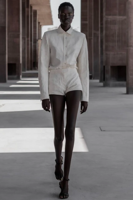 A fashionable dark-skinned woman in a linen suit walks toward the camera in a light and airy outdoor architectural space.