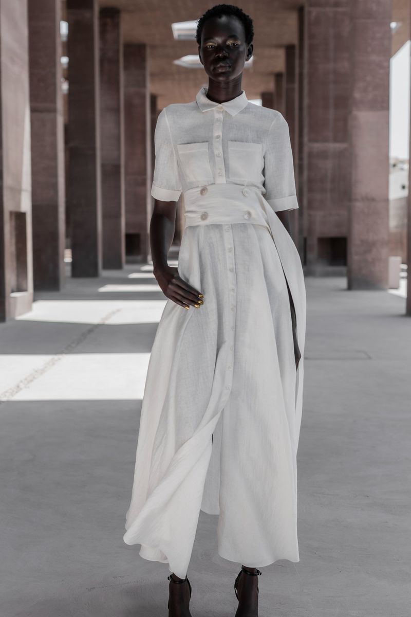 An African woman in a white dress stands confidently in an outdoor colonnade featuring concrete columns on a sunny day for a fashion shot.