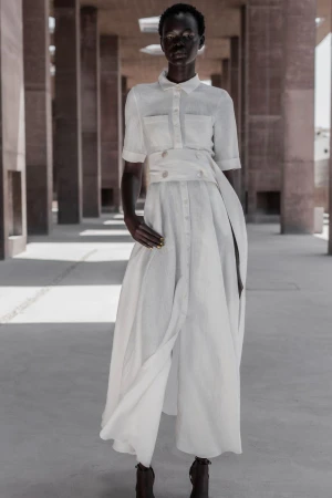 An African woman in a white dress stands confidently in an outdoor colonnade featuring concrete columns on a sunny day for a fashion shot.