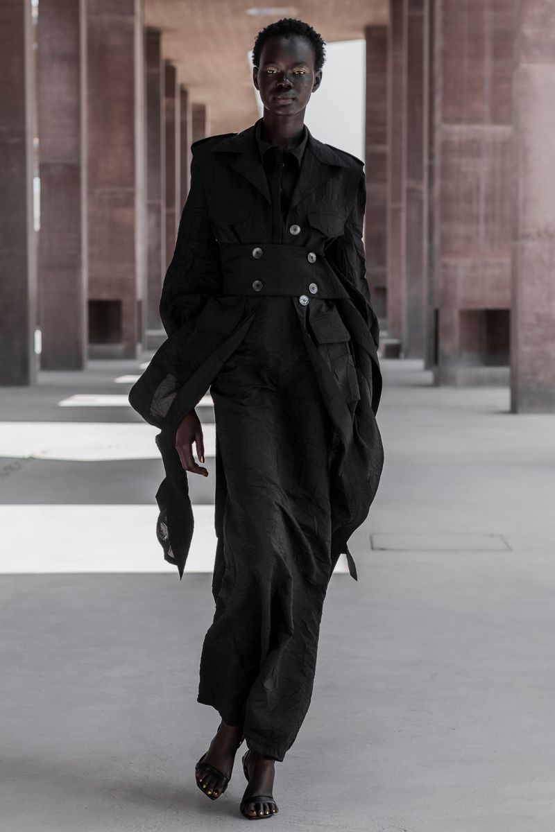 Runway-style shot of a model wearing all-black clothing in a concrete hallway with pillars. The model is wearing a coat with a corset.