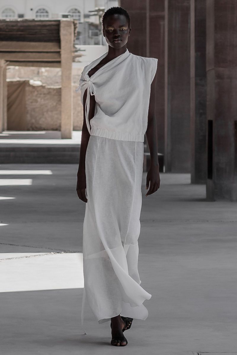 A dark-skinned woman models white clothing in a sunlit outdoor architectural setting, creating a high-fashion editorial image.