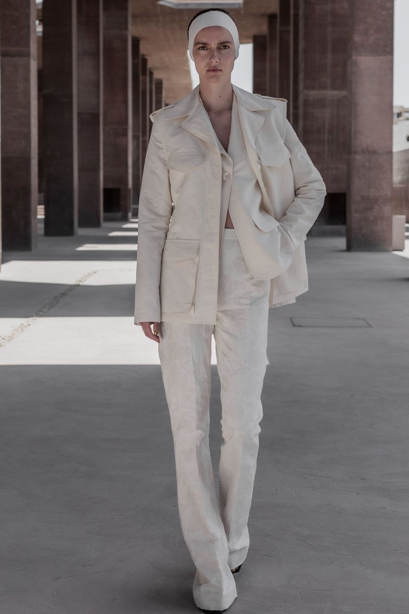 Woman in runway fashion posing in a formal white pantsuit with a matching head wrap in a concrete outdoor setting with pillars.
