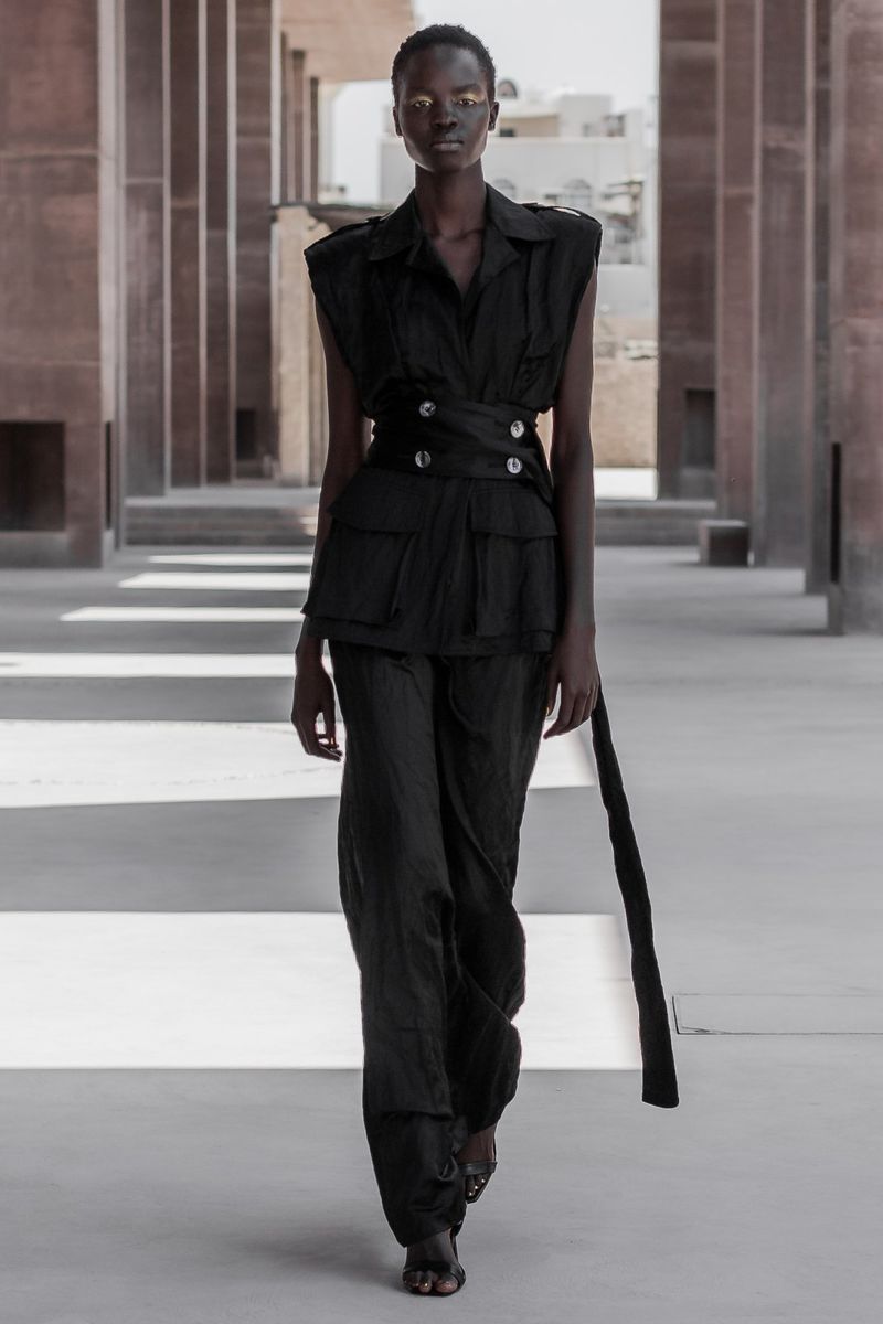 Runway model in a black, sleeveless suit with gold eye makeup struts down the catwalk in front of a brick-columned backdrop.
