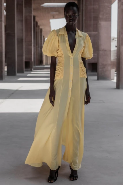 A Black woman in a pastel yellow dress stands in a covered outdoor space with concrete columns, showcasing fashion and style.