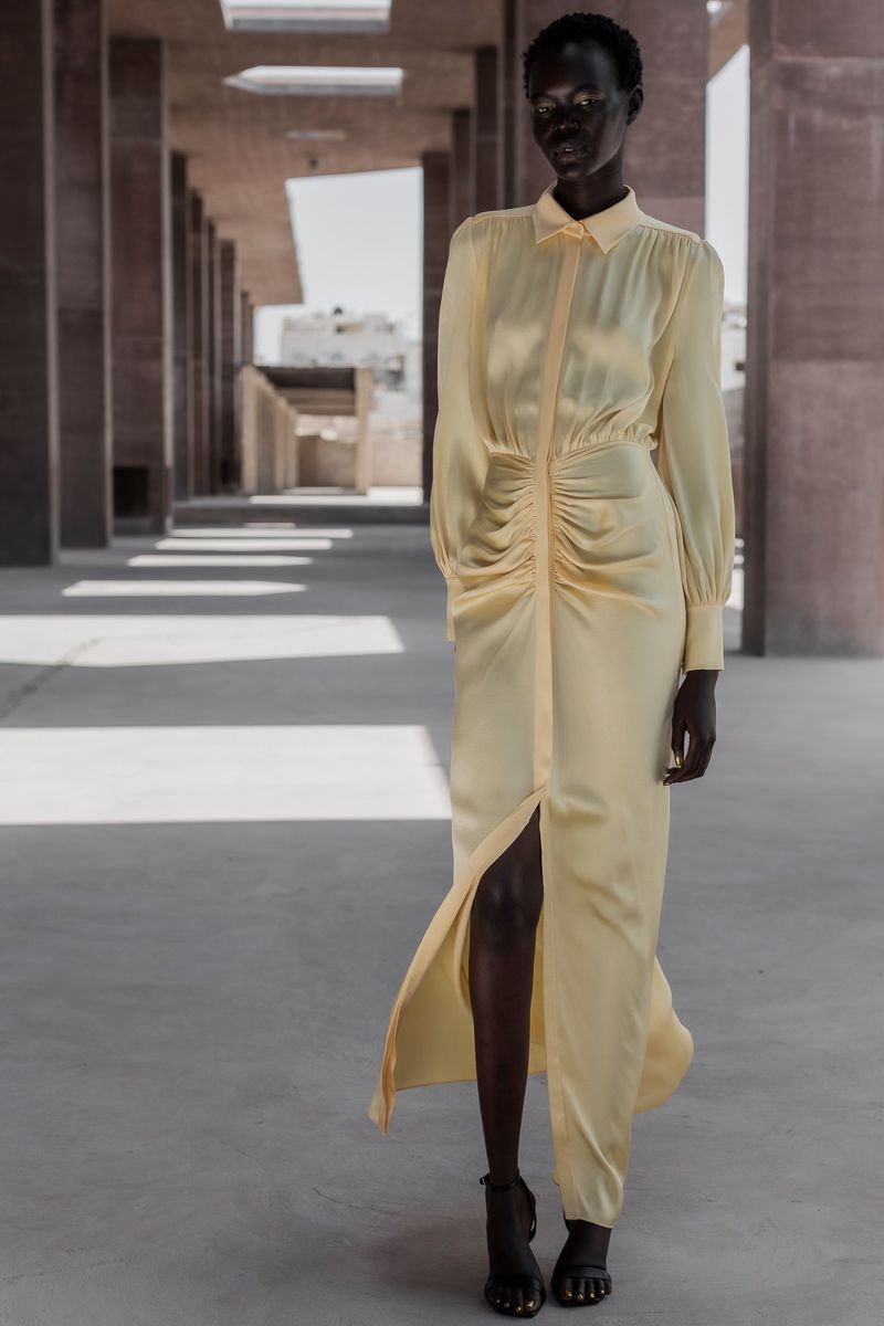 Woman in a pale yellow, long-sleeved dress with a front slit poses in an outdoor corridor with concrete columns and floors.