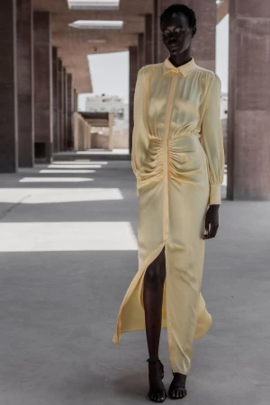 Woman in a pale yellow, long-sleeved dress with a front slit poses in an outdoor corridor with concrete columns and floors.