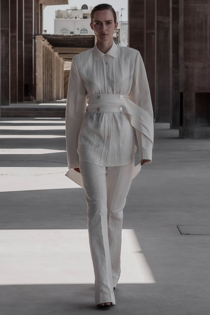 A woman in a white linen outfit walks toward the camera, wearing an oversized belted shirt and wide-leg pants, with architectural backdrop.