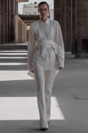 A woman in a white linen outfit walks toward the camera, wearing an oversized belted shirt and wide-leg pants, with architectural backdrop.