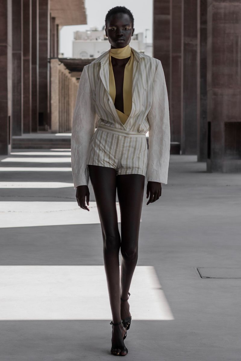 Fashion model walking outdoors in a striped linen shorts suit with a yellow scarf against an architectural background in natural light.