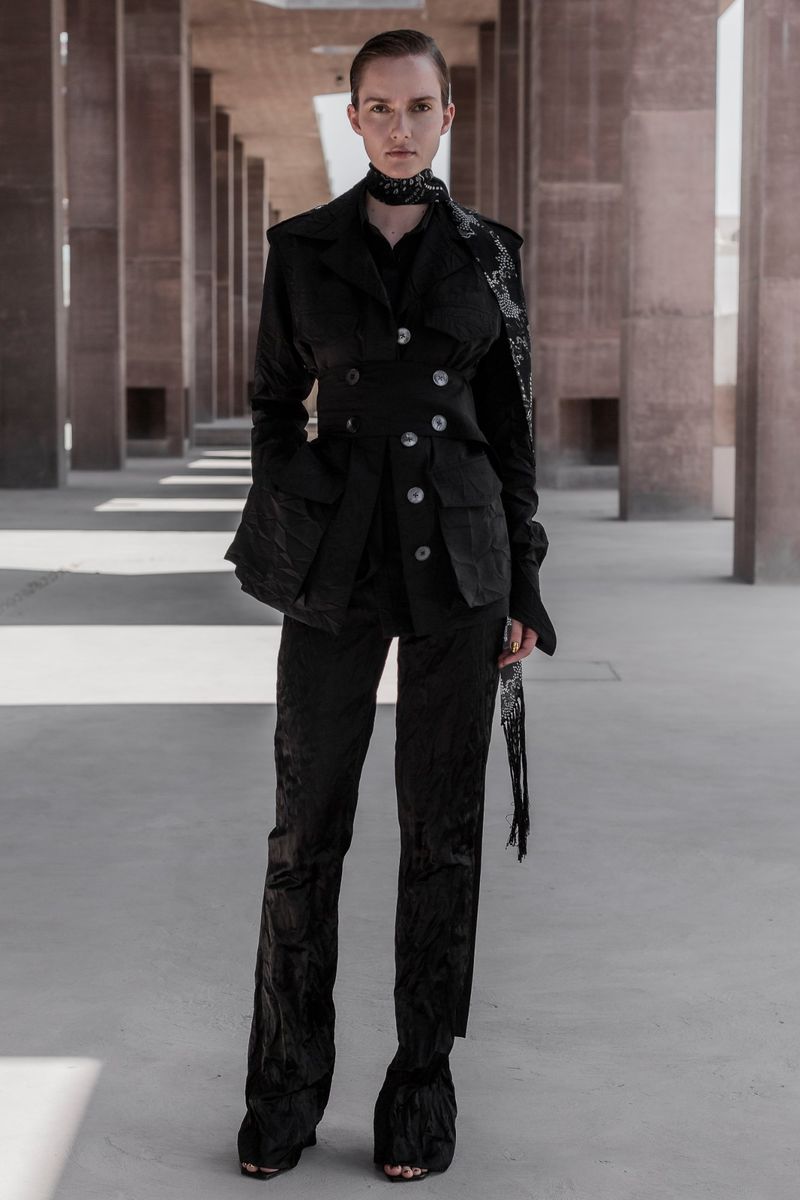 A woman in a black textured suit poses outdoors in an architectural space, complemented by a matching scarf and shoes, under neutral lighting.