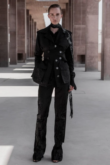 A woman in a black textured suit poses outdoors in an architectural space, complemented by a matching scarf and shoes, under neutral lighting.