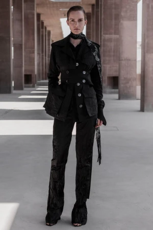 A woman in a black textured suit poses outdoors in an architectural space, complemented by a matching scarf and shoes, under neutral lighting.