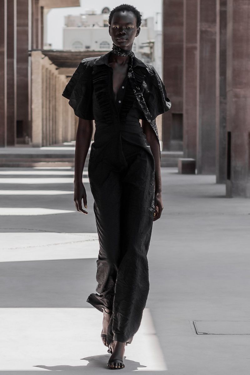 A dark-skinned adult woman walks in a black jumpsuit, with columns behind her. Fashion editorial photography, full-length shot.