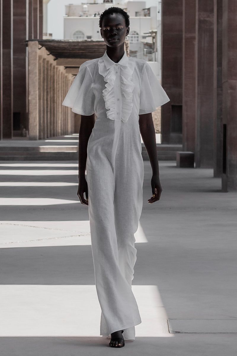 An adult woman in a white jumpsuit with ruffles poses in a covered outdoor space with light and shadows from a colonnade.