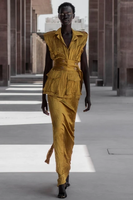 Woman in a golden-yellow two-piece outfit with a layered top and a long skirt, posing in a corridor with pillars and sunlight.