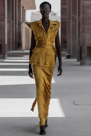 Woman in a golden-yellow two-piece outfit with a layered top and a long skirt, posing in a corridor with pillars and sunlight.