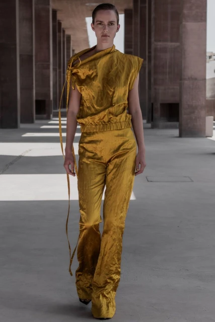 Fashion model wears a gold crushed-velvet-like top and pants walking in an outdoor colonnade with concrete columns and neutral light.