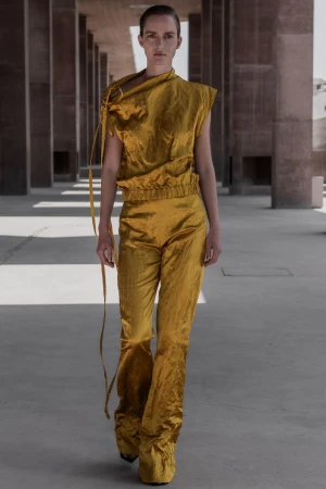 Fashion model wears a gold crushed-velvet-like top and pants walking in an outdoor colonnade with concrete columns and neutral light.
