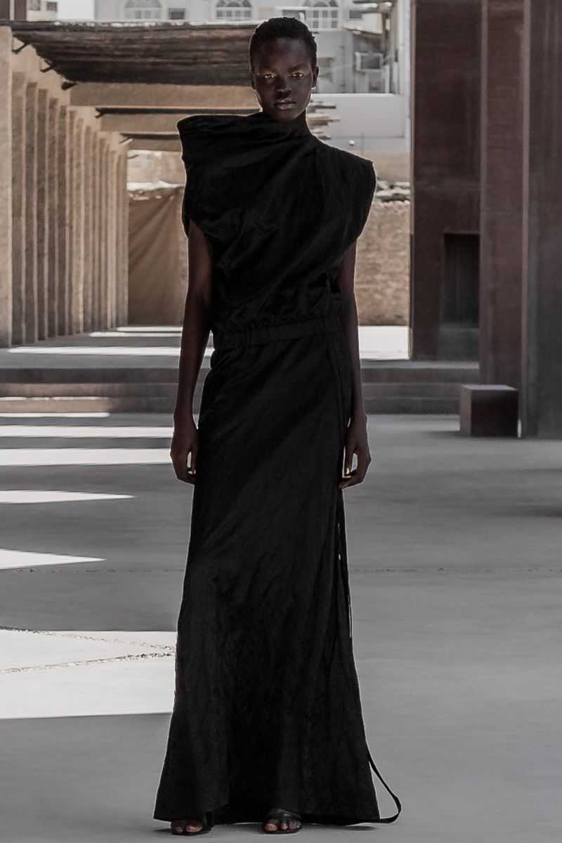 A solo Black fashion model poses in a dark dress with natural light highlighting the architecture and textures of an outdoor structure.