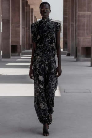 Model Akolde Meen walking forward in a printed black high-neck sleeveless top and matching trousers through a concrete colonnade.