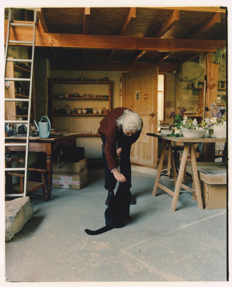 Woman with grey hair bending down to pet a black cat on the floor inside a rustic pottery studio with wooden shelves.