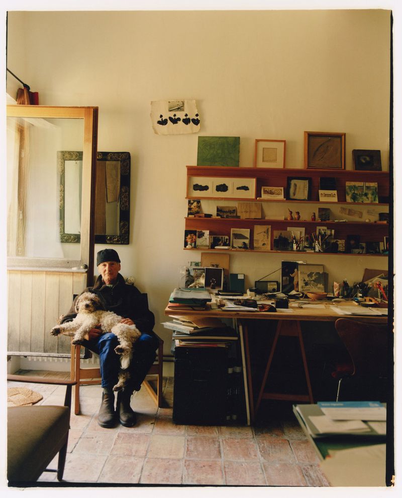 Man seated on a wooden chair holding a fluffy dog on his lap in an artist studio with shelves and a wooden drafting desk.
