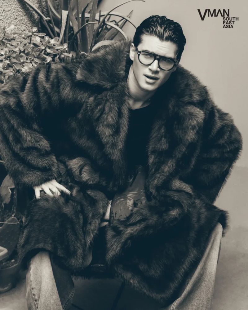 Man wearing heavy faux fur coat and thick black eyeglasses seated outdoors in monochrome portrait