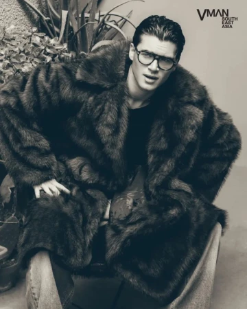 Man wearing heavy faux fur coat and thick black eyeglasses seated outdoors in monochrome portrait