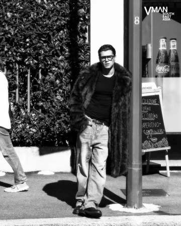 Antonio Medugno wearing a long fur coat, black shirt, and baggy jeans leaning against a post on a city street in black and white.