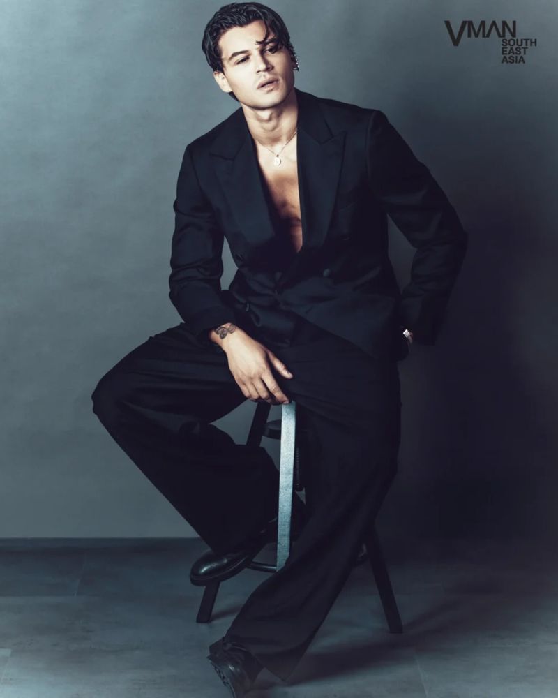 Antonio Medugno seated on a stool wearing a black tailored suit with an open jacket against a grey studio backdrop.