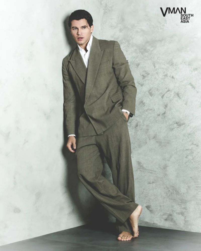 Man wearing an olive suit leaning against a grey concrete corner in a V MAN SEA editorial photograph