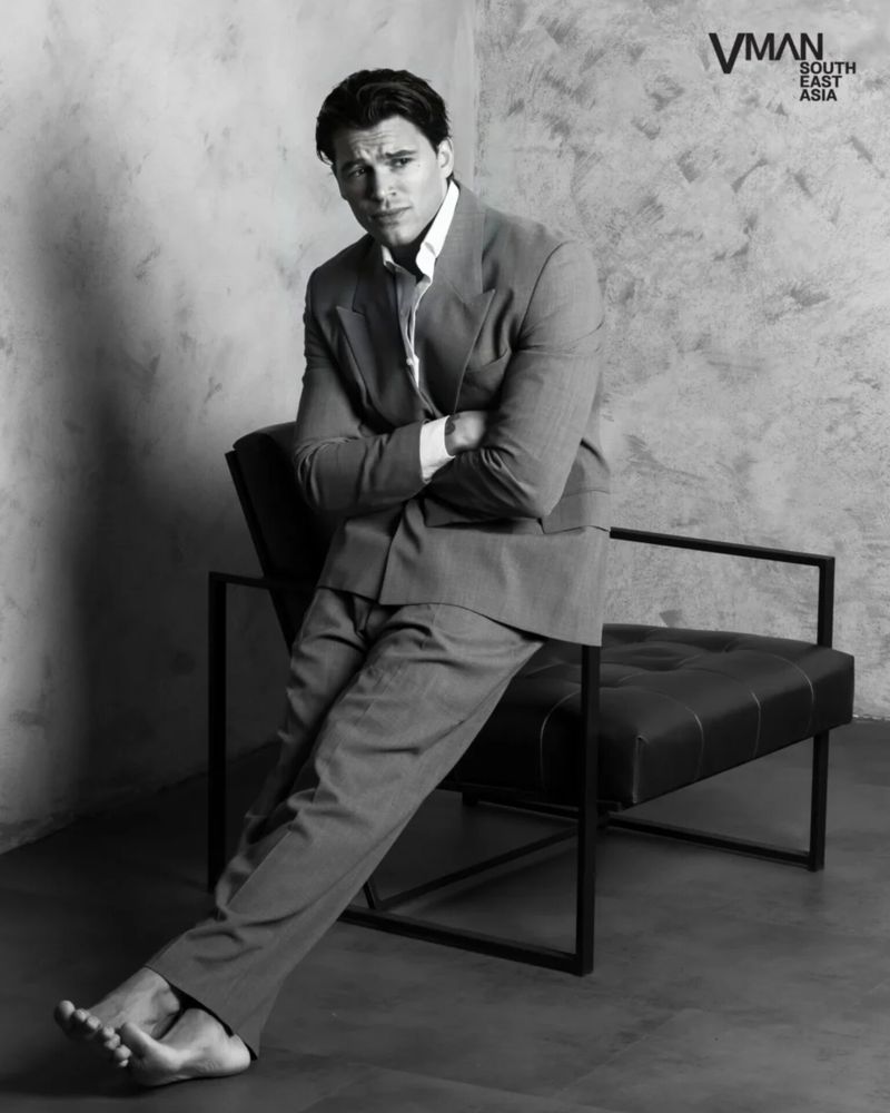 Barefoot man in a tailored suit leaning on a modern black leather chair against a textured studio wall.