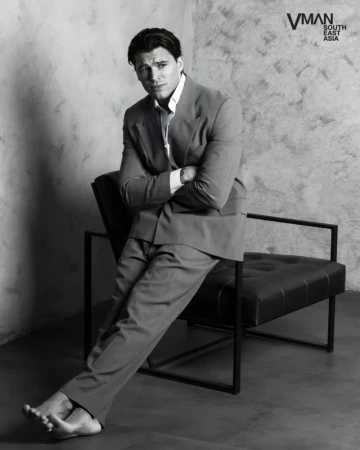 Barefoot man in a tailored suit leaning on a modern black leather chair against a textured studio wall.