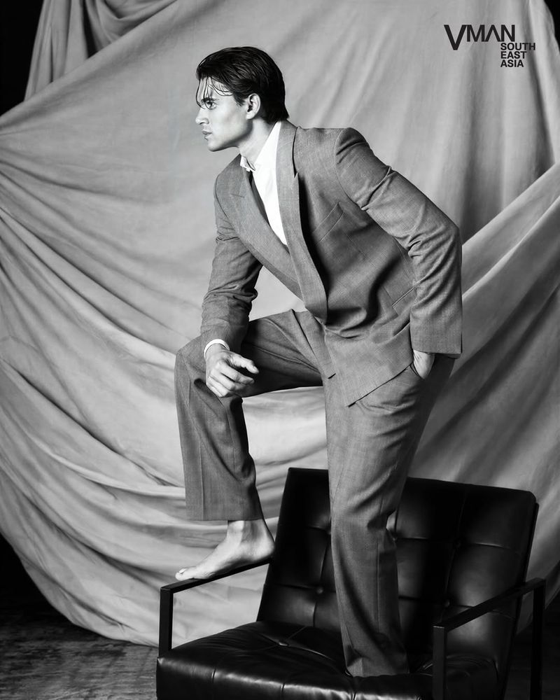 Antonio Medugno posing barefoot on a black leather armchair wearing a tailored suit against draped fabric in black and white.