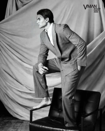 Antonio Medugno posing barefoot on a black leather armchair wearing a tailored suit against draped fabric in black and white.