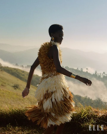 Model wearing a textured tan and cream feathered dress standing on a grassy hillside with misty rolling mountains.