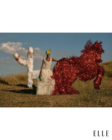 Model in a white dress standing in a grassy field next to large red and white tinsel sculptures under a blue sky.