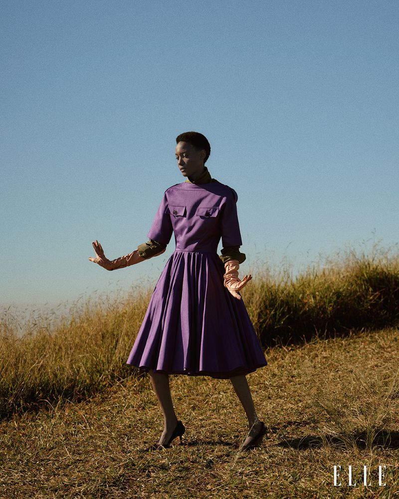 Model wearing a purple pleated midi dress and long gloves poses in a dry grassy field under a clear blue sky.