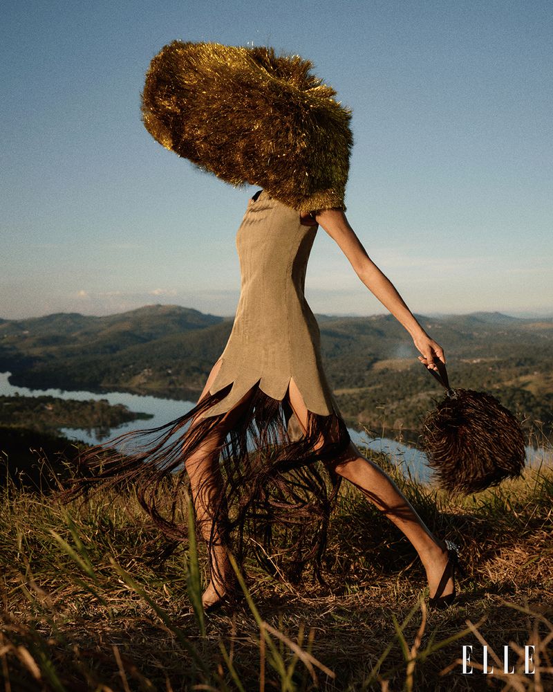 Model wearing an olive fringe dress and an oversized gold tinsel headpiece walking on a grassy hill overlooking a lake for Elle Brasil.