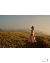 Model standing in a grassy field surrounded by mist, wearing a long flowing pink gown in warm morning light.