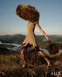 Model wearing an olive fringe dress and an oversized gold tinsel headpiece walking on a grassy hill overlooking a lake for Elle Brasil.