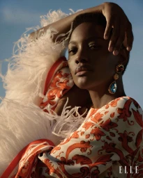 Model wearing a patterned orange and white dress with feather trim against a clear blue sky