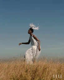 Model wearing a textured white fringe dress and a matching feather headdress standing in a dry golden grass field under a clear blue sky.
