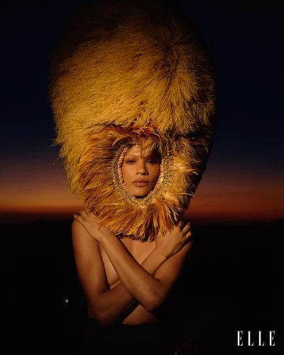 Model wearing an oversized golden feather headdress with arms crossed against a dark twilight sunset sky.