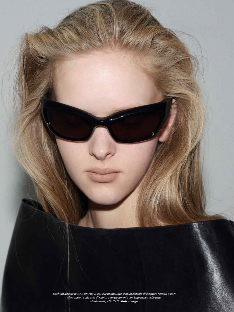 Close-up portrait of a blonde woman wearing dark angular sunglasses and a structured black leather high collar against a grey background.