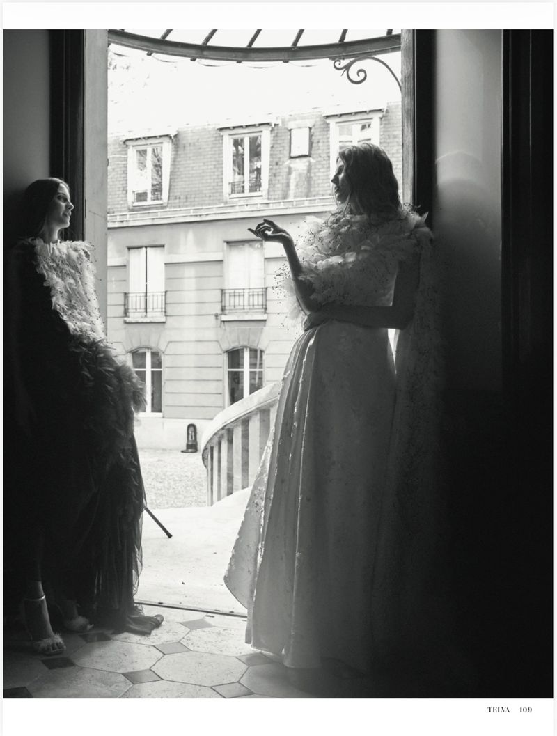 Black and white editorial photo of two models wearing feathered gowns standing in an open doorway overlooking a courtyard.