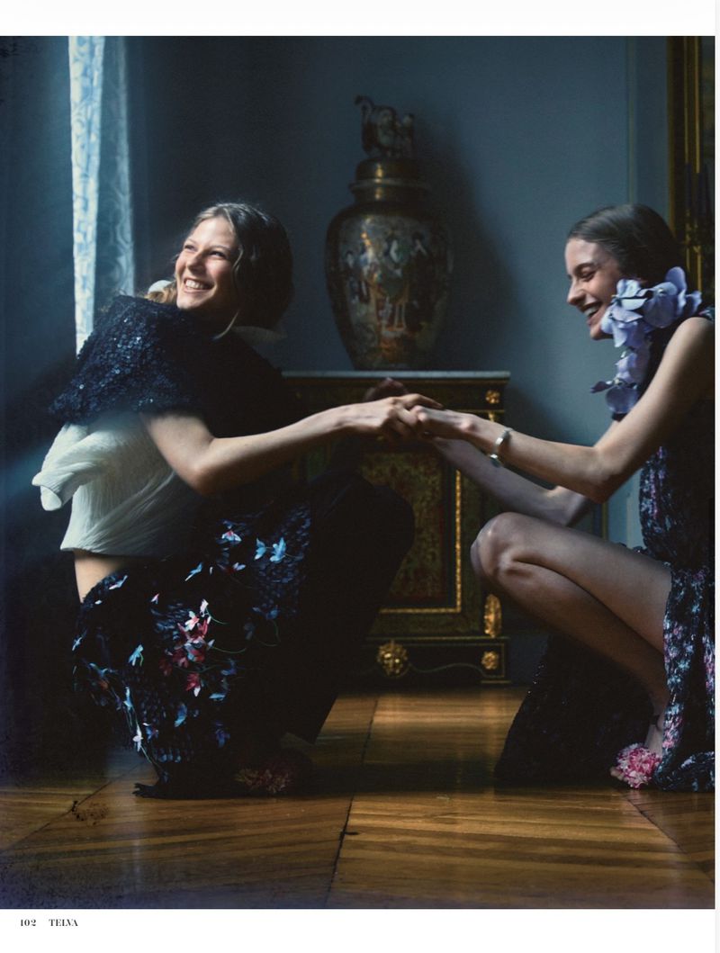 Two smiling female models squatting indoors on wooden floors, holding hands and laughing in a dimly lit room.