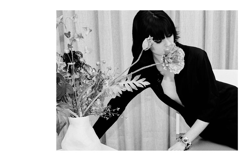 Black and white editorial image of a model leaning over a vase of flowers with a white backdrop.