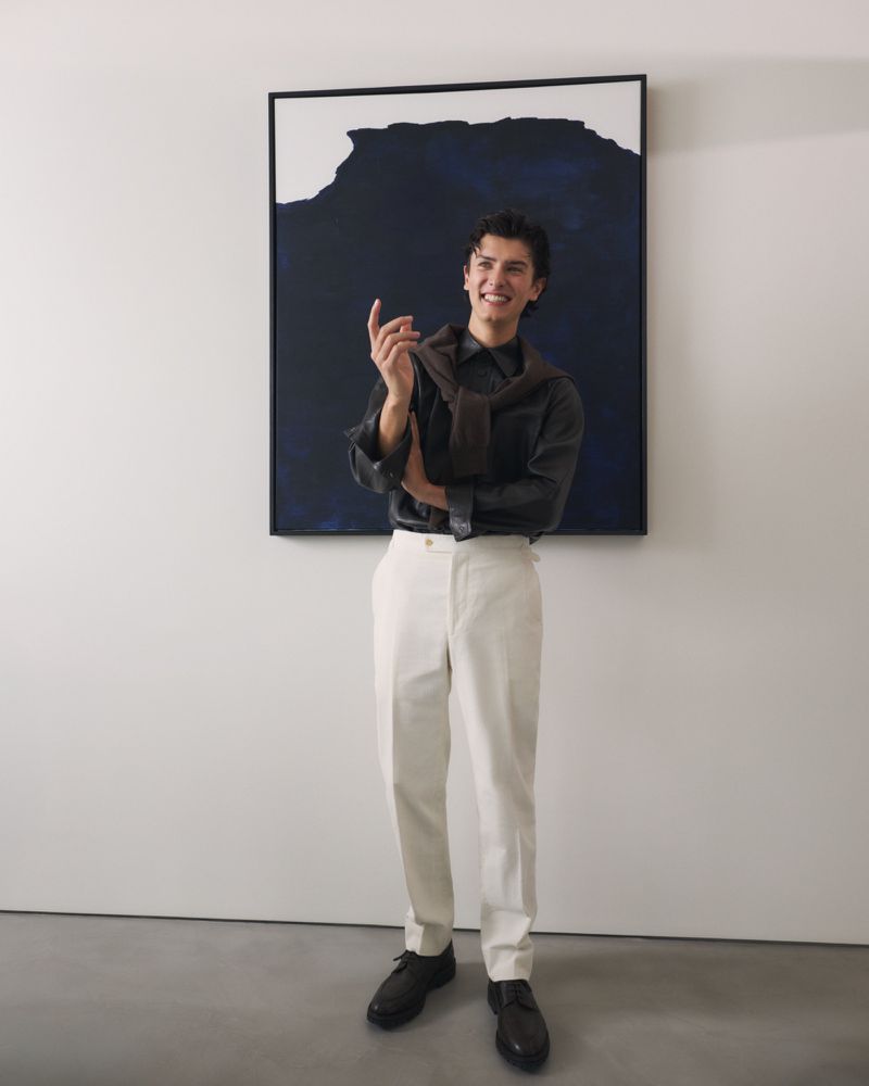 Count Nikolai of Monpezat smiling in white trousers and a dark shirt with a draped sweater, standing in front of an abstract painting.