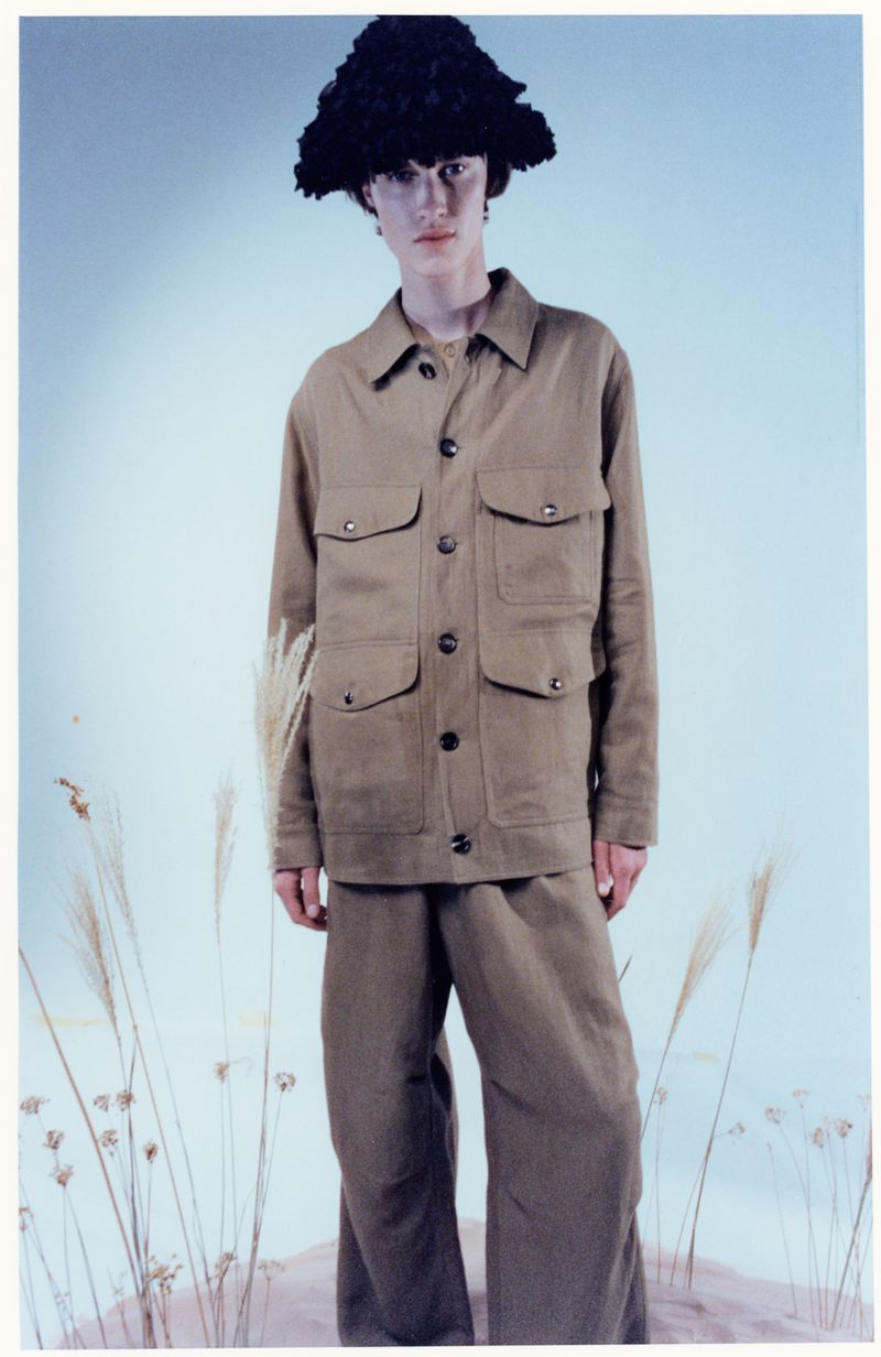 Model Boy Dijkhuis wears a khaki utility suit and textured black hat amidst dried reeds in a studio editorial.
